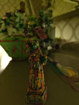 Small vertical mixed-media sculpture covered in layered pink, green, yellow and dark paint, with a tiny raised element at its top