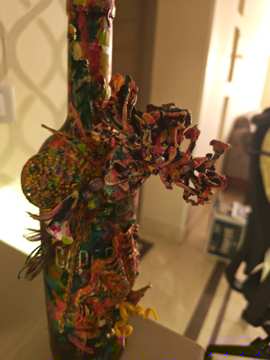 Mixed-media glass bottle sculpture covered in vivid layered colours, textured attachments, branches and small sculptural forms